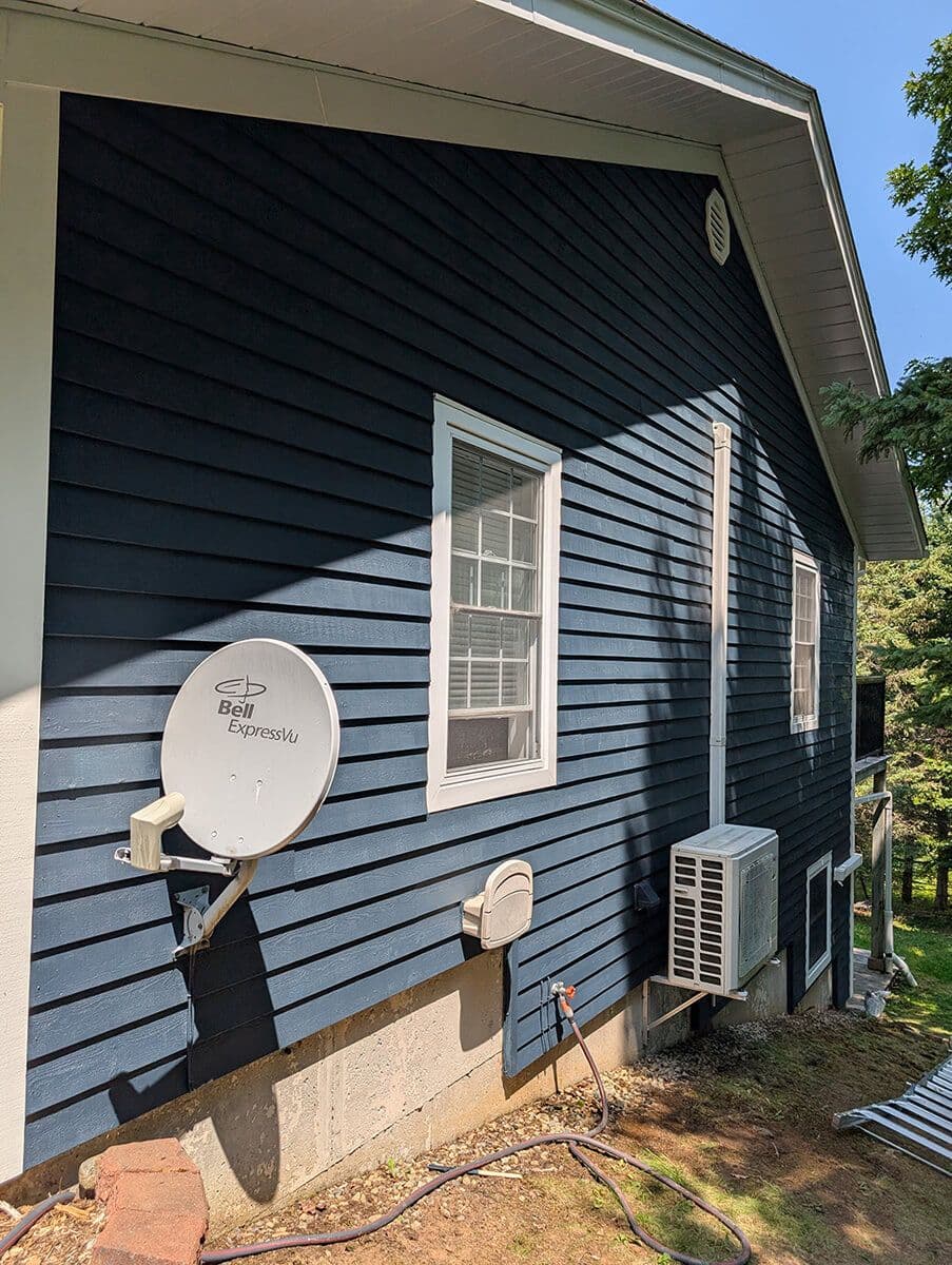 House side with dark blue horizontal siding, white trim, satellite dish, and air conditioning unit.