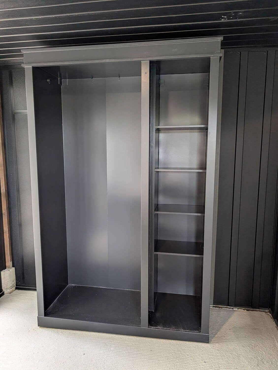 Empty black wardrobe with hanging space and built-in shelving against dark paneled wall.