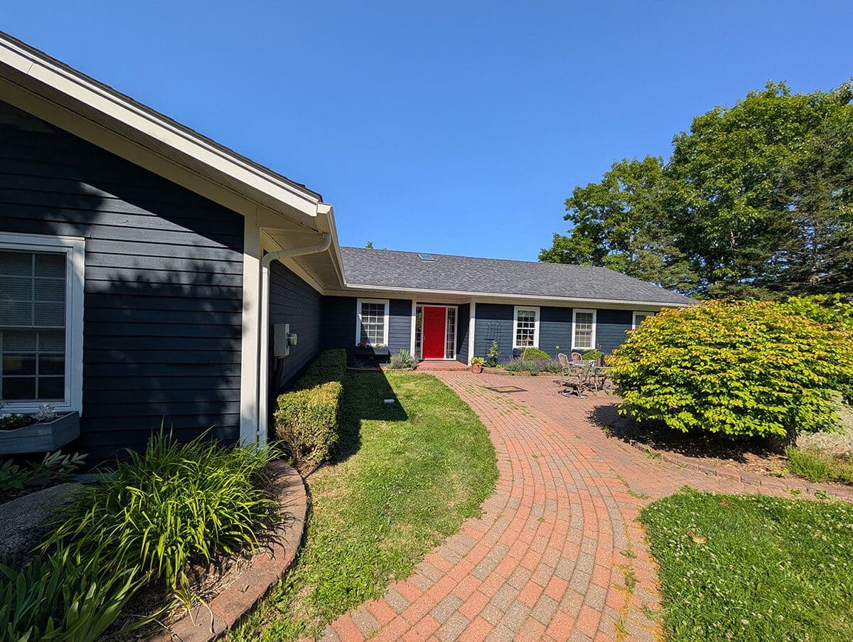 Brick walkway leading to a blue siding house with red front door, white trim, and landscaped yard.
