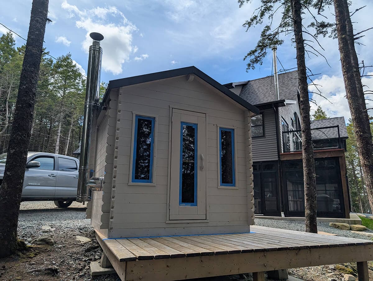 Small light-colored wooden cabin with steel chimney pipe and narrow windows on raised deck in wooded setting.